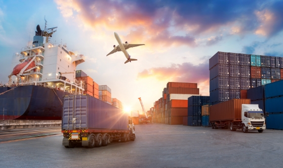 A busy shipping yard at sunset, featuring a large cargo ship docked on the left, a stack of colorful shipping containers, a semi-truck transporting a container, and an airplane flying overhead. The sky displays vibrant colors of the setting sun.